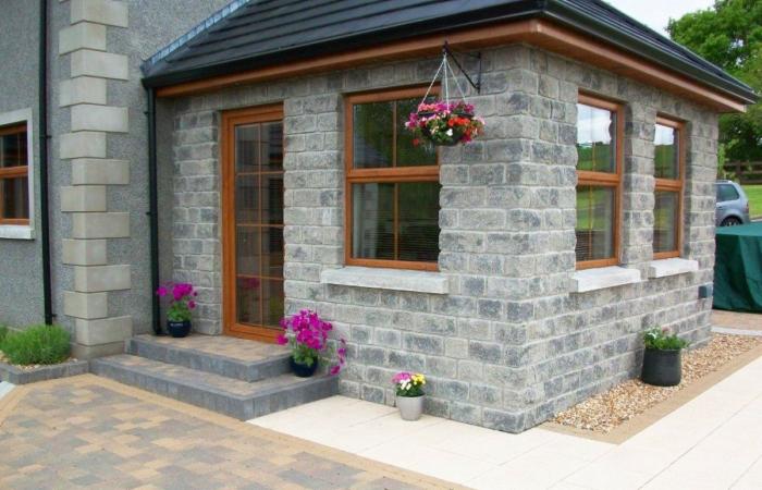 Kamienna elewacja domu z drewnianymi oknami i drzwiami. Brukowany chodnik prowadzi do wejścia. Kwiaty w wiszącej donicy i doniczkach dodają uroku.