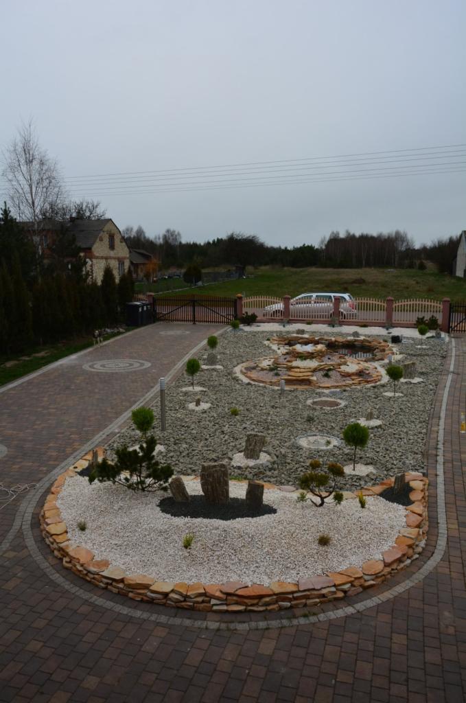 Ogród z oczkiem wodnym i kamiennymi elementami dekoracyjnymi, widok z góry. Brązowa kostka brukowa i ogrodzenie w tle. Pochmurne niebo.