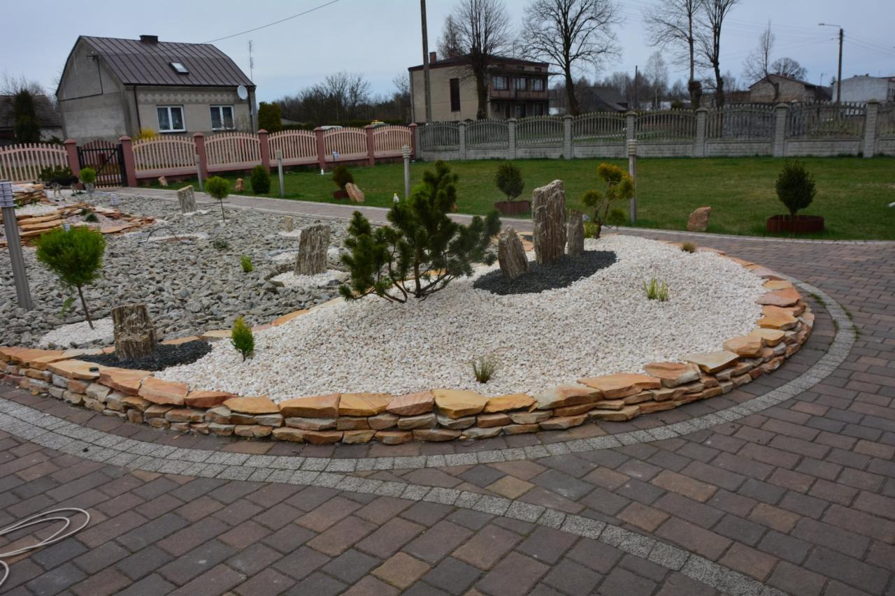 Ogród z kamieniami, niską roślinnością i obrzeżem z piaskowca, otoczony brukowaną ścieżką. W tle domy i ogrodzenia.