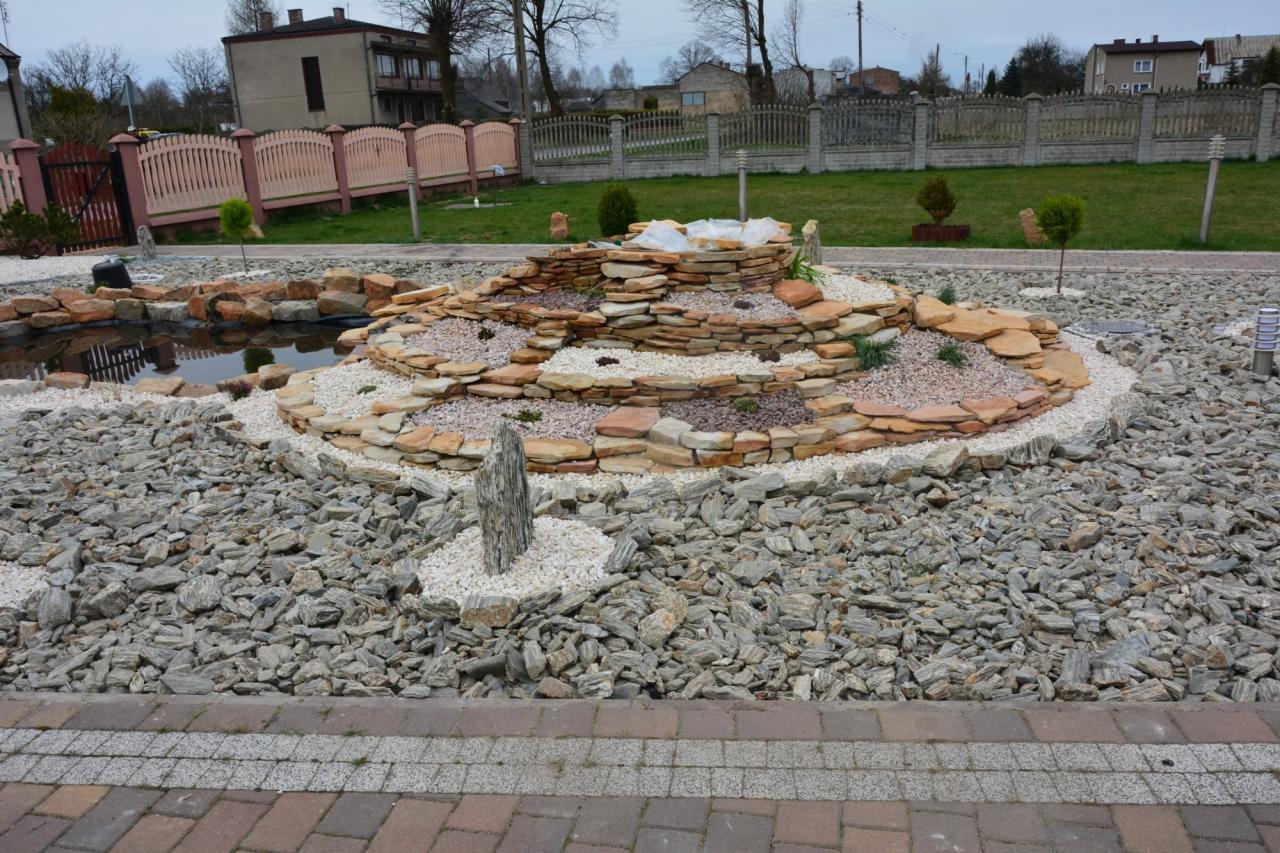 Ogród z oczkiem wodnym i kaskadą z kamienia, otoczony szarym żwirem i ozdobnymi roślinami. W tle budynki i ogrodzenia.