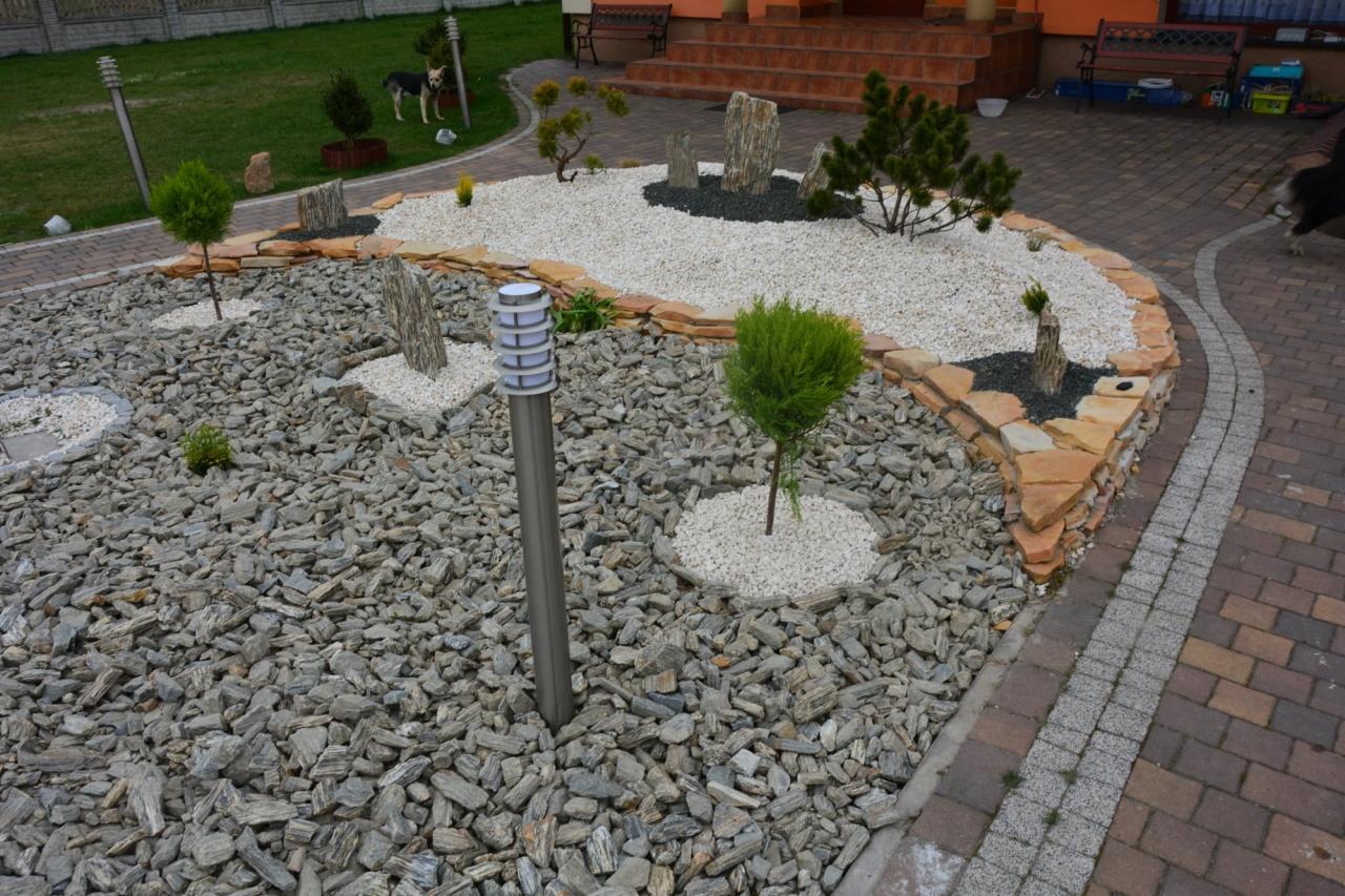 Ogród w Blachowni z kamienną rabatą, ozdobnymi kamieniami, niskimi drzewkami i oświetleniem. Estetyczne wykończenie obrzeży kostką brukową.