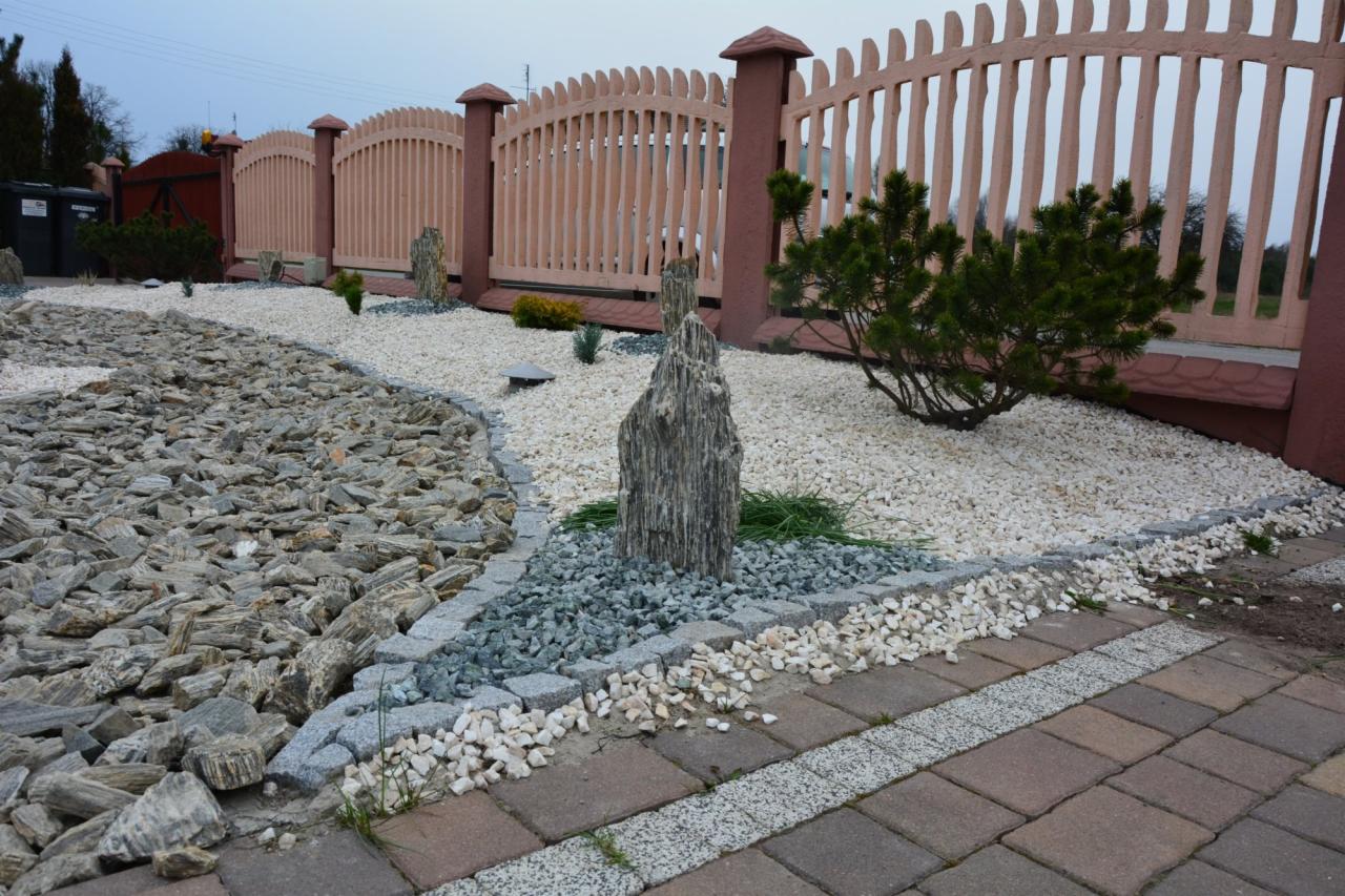 Ogród z dekoracyjnym kamieniem, ścieżką z łupka i niską, beżową palisadą. Białe i szare kamienie tworzą rabaty, a w tle widoczna zielona roślinność.