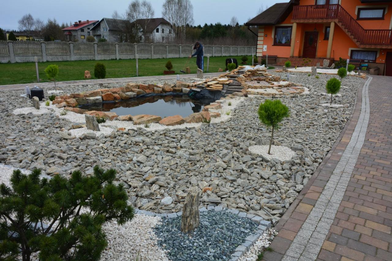 Ogród z oczkiem wodnym otoczonym kamieniami i wysypany żwirem, z młodymi drzewkami i elementami dekoracyjnymi. W tle dom z pomarańczową elewacją i zielony trawnik.