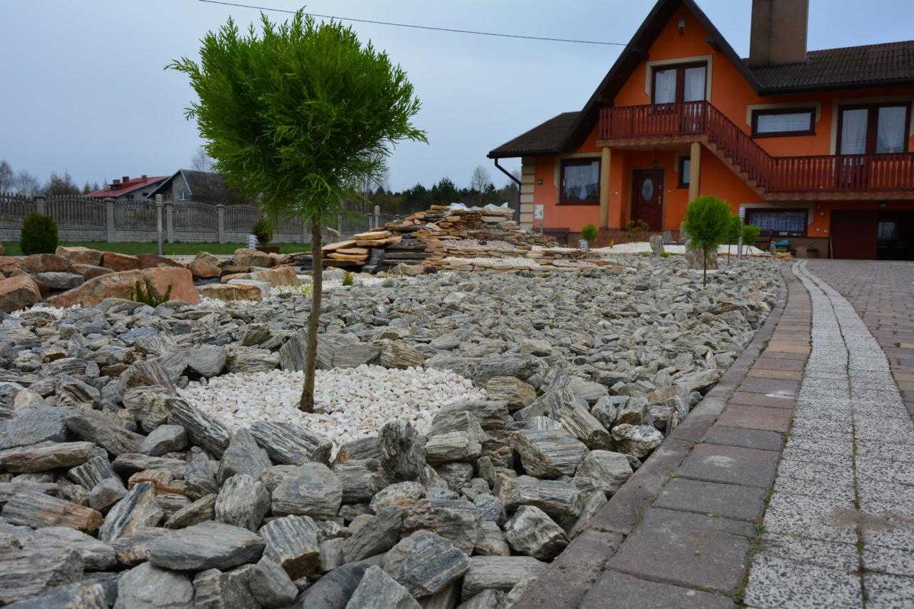 Ogród z drzewkiem bonsai, kamieniami ozdobnymi i ścieżką z kostki brukowej prowadzącą do pomarańczowego domu z balkonem. Widok na starannie zaprojektowany teren.
