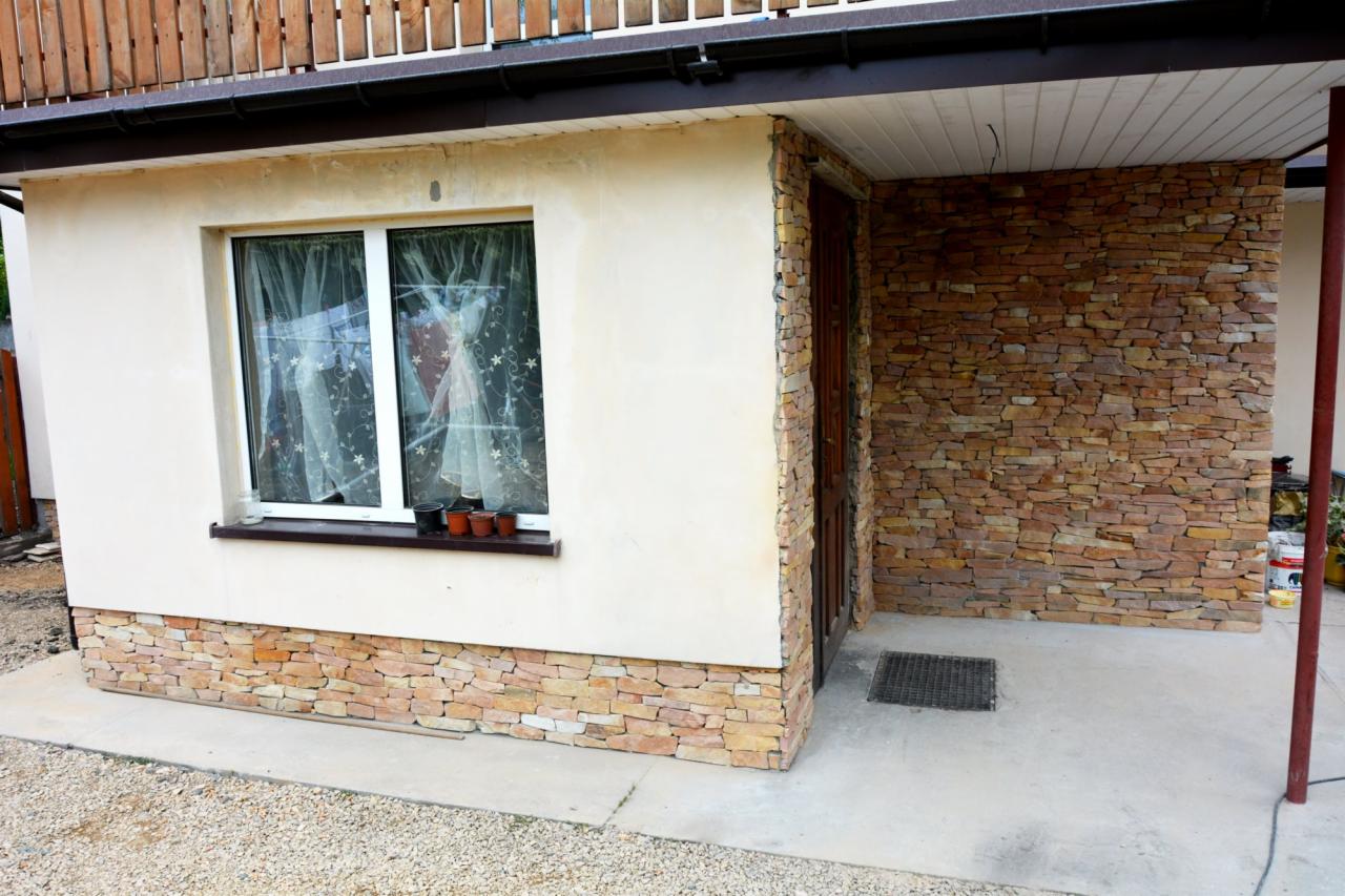 Wykończenie elewacji kamieniem dekoracyjnym wokół wejścia i u podstawy ściany budynku. Kremowy tynk kontrastuje z ciepłą barwą kamienia.