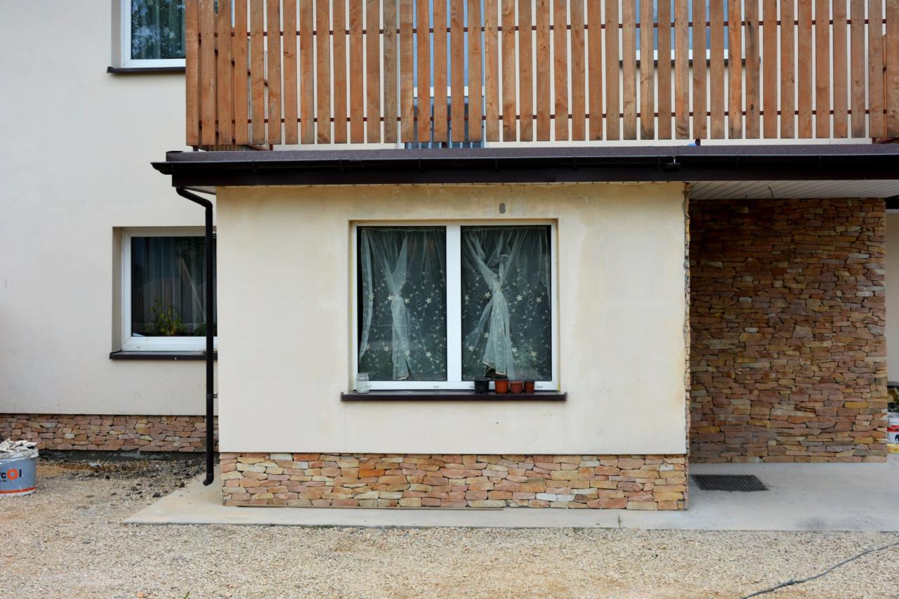 Elewacja budynku z drewnianą balustradą balkonu i kamienną okładziną. Widoczna okładzina z kamienia elewacyjnego na cokole i ścianie wnęki wejściowej.