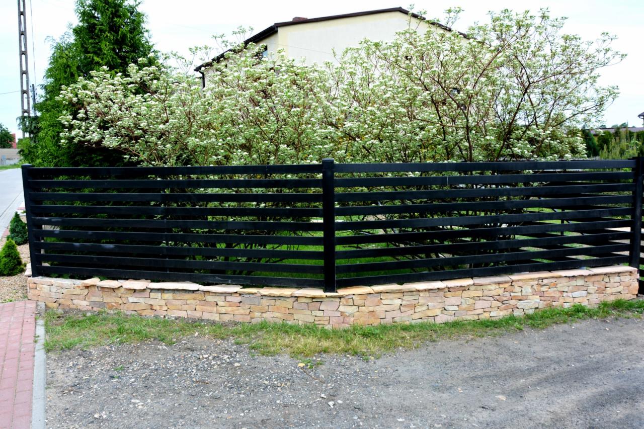 Nowoczesne ogrodzenie z czarnych, poziomych desek na kamiennej podmurówce. W tle kwitnące krzewy i dom. Ujęcie z zewnątrz posesji, widoczny fragment chodnika i podjazdu.