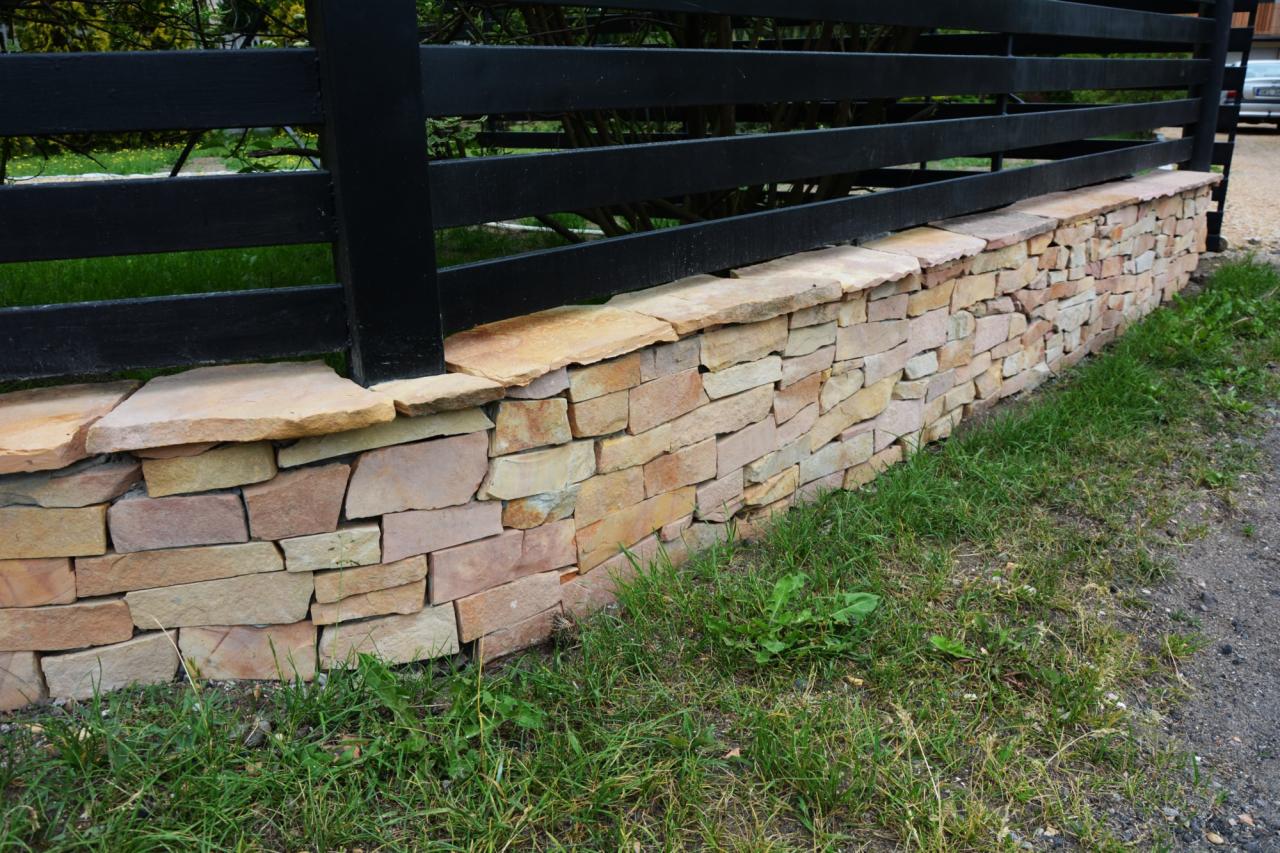 Kamienna podmurówka ogrodzenia z czarnymi, drewnianymi przęsłami. Niska ściana z nieregularnych, piaskowych kamieni, obok trawa i ścieżka.