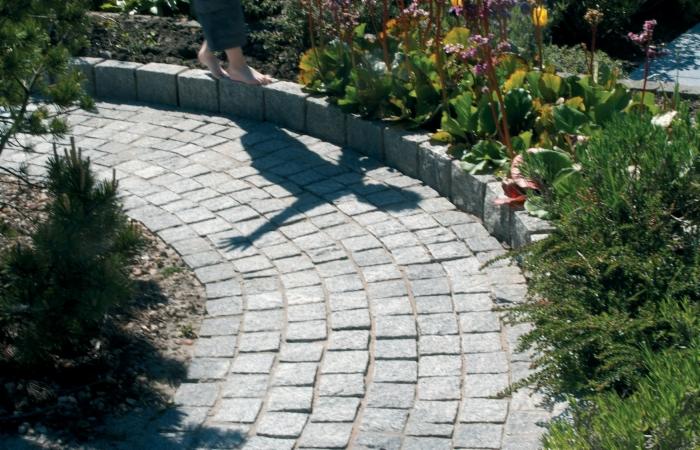 Zakrzywiony chodnik z szarej kostki, obramowany kamieniami. Cień osoby przechodzącej po obrzeżu. Roślinność wzdłuż ścieżki.