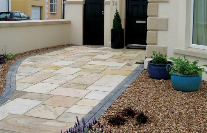 Jasna, kamienna ścieżka prowadzi do domu z czarnymi drzwiami. Brzegi wykończone szarą kostką i żwirem. Donice z roślinami przy wejściu. Numer domu 74.