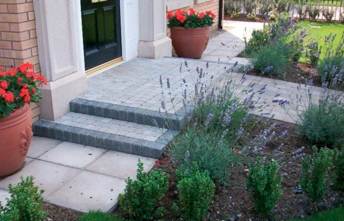 Ułożona kostka brukowa tworzy schody wejściowe i ścieżkę w ogrodzie, z donicami z czerwonymi kwiatami i lawendą. Detal wykończenia.