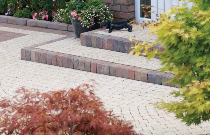 Ułożona kostka brukowa w jasnych odcieniach z obrzeżami z ciemniejszej kostki. Na schodach figurka jamnika, obok rośliny ozdobne.