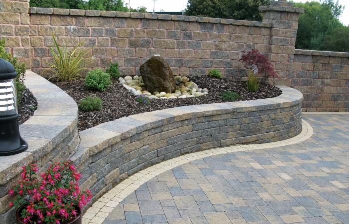 Ogród z fontanną z kamienia, obrzeżem z kostki i podwyższonymi rabatami otoczonymi murem z kamienia. Kostka brukowa tworzy wzór na podjeździe.