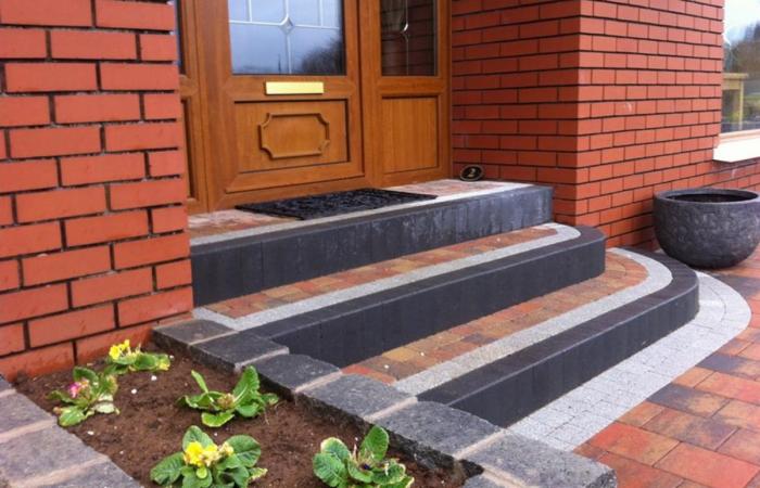Kaskadowe schody wejściowe z kostki brukowej w odcieniach szarości i cegły, prowadzące do drewnianych drzwi. Uroku dodaje rabata z kwiatami i donica.
