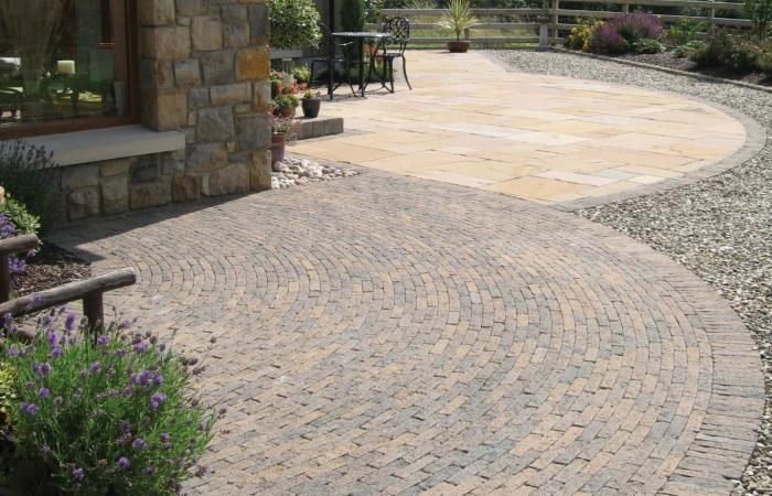 Wykończony taras z kostki brukowej ułożonej w łuk, z elementami kamiennymi i żwirowym obrzeżem. Widoczny fragment budynku z kamienia i roślinność.