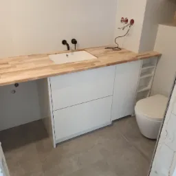 Łazienka w trakcie remontu: białe szafki pod umywalką z drewnianym blatem, czarne baterie, widoczne kable elektryczne i toaleta z boku.