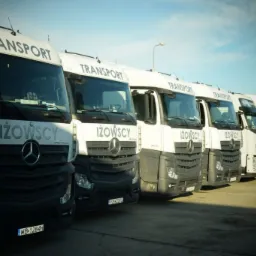 Flota białych ciężarówek Mercedes-Benz firmy Iżowscy Transport, ustawionych w rzędzie, z widocznymi numerami rejestracyjnymi i logo firmy.