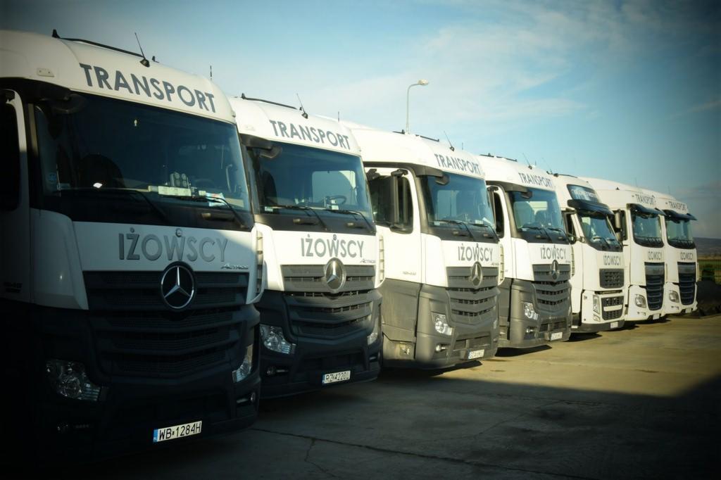 Flota białych ciężarówek Mercedes-Benz firmy Iżowscy Transport, ustawionych w rzędzie, z widocznymi numerami rejestracyjnymi i logo firmy.
