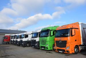 Rząd ciężarówek Mercedes-Benz w różnych kolorach (biały, zielony, pomarańczowy) zaparkowanych przed budynkiem w słoneczny dzień. Widoczne błękitne niebo z chmurami.