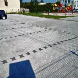 Parking z szarej kostki brukowej z czarnymi wstawkami wyznaczającymi miejsca postojowe, w tle budynek i kolorowy plac zabaw.