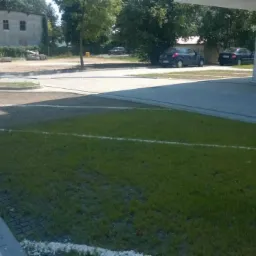 Wykończony parking z ażurowymi płytami trawnikowymi i kostką brukową, z białymi liniami oddzielającymi miejsca postojowe, w tle budynek i zaparkowane samochody.