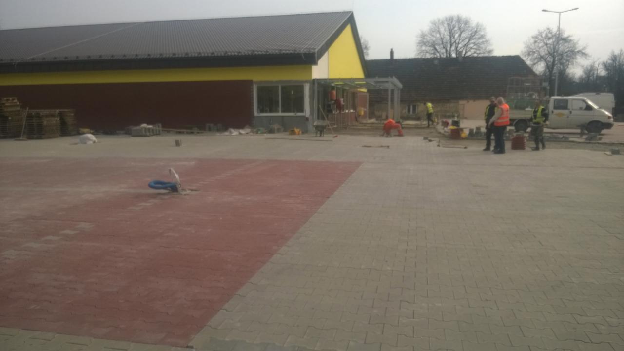 Układanie dwukolorowej kostki brukowej na placu przed budynkiem. Ekipa w kamizelkach odblaskowych pracuje, widoczne palety z kostką i samochód dostawczy.