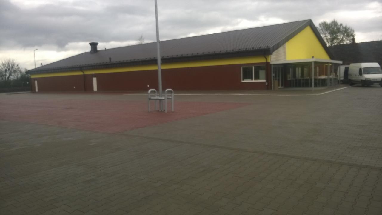 Rozległy parking z kostki brukowej przed budynkiem w odcieniach brązu i żółci, w tle zaparkowane busy dostawcze, pochmurne niebo.