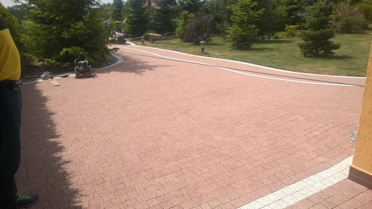 Czerwona kostka brukowa ułożona wzdłuż ścieżki z białym obramowaniem na posesji. W tle zieleń drzew i krzewów, widoczna maszyna do ubijania kostki.