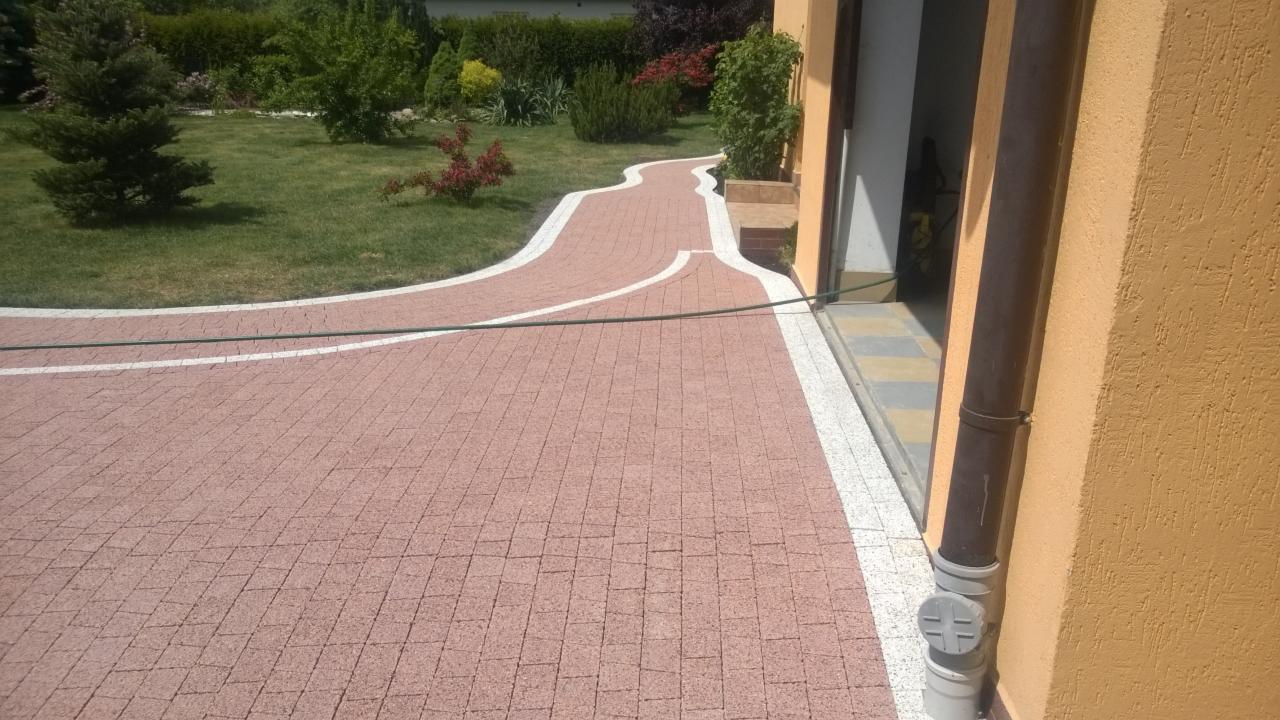 Kremowo-różowa kostka brukowa z białymi obrzeżami tworzy wijącą się ścieżkę przez trawnik, prowadzącą do wejścia budynku z żółtą ścianą i brązową rynną.