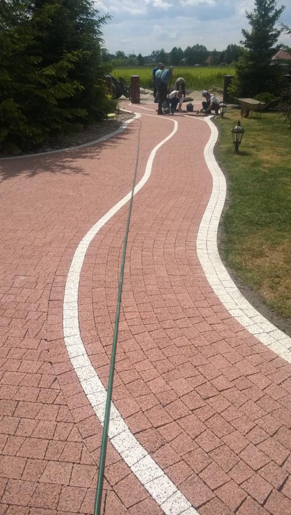 Kaskadowa ścieżka z czerwonej kostki brukowej z białymi obrzeżami, w tle grupa robotników podczas układania, otoczona zielenią. Widok z góry, lekko z ukosa.