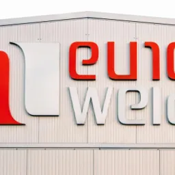 Zewnętrzny szyld firmy Euro Weld z czerwono-białym logo na tle białej, blaszanej elewacji budynku.