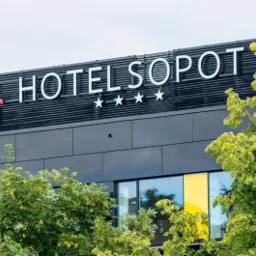 Elewacja budynku hotelu Sopot z podświetlanym napisem i logo z trzema gwiazdkami, widoczna zza zielonych drzew.