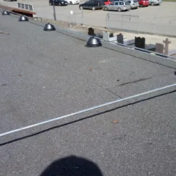 Instalacja odgromowa na płaskim dachu pokrytym papą, z widocznymi czarnymi podstawami i metalowymi przewodami, w tle nagrobki i parking.