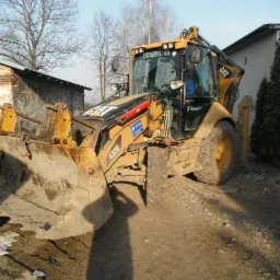 Żółta koparko-ładowarka Caterpillar 432E stoi na nierównym terenie z gruzem, obok ceglanego budynku i wysokich tuj, widoczne ślady użytkowania i zabrudzenia na maszynie.