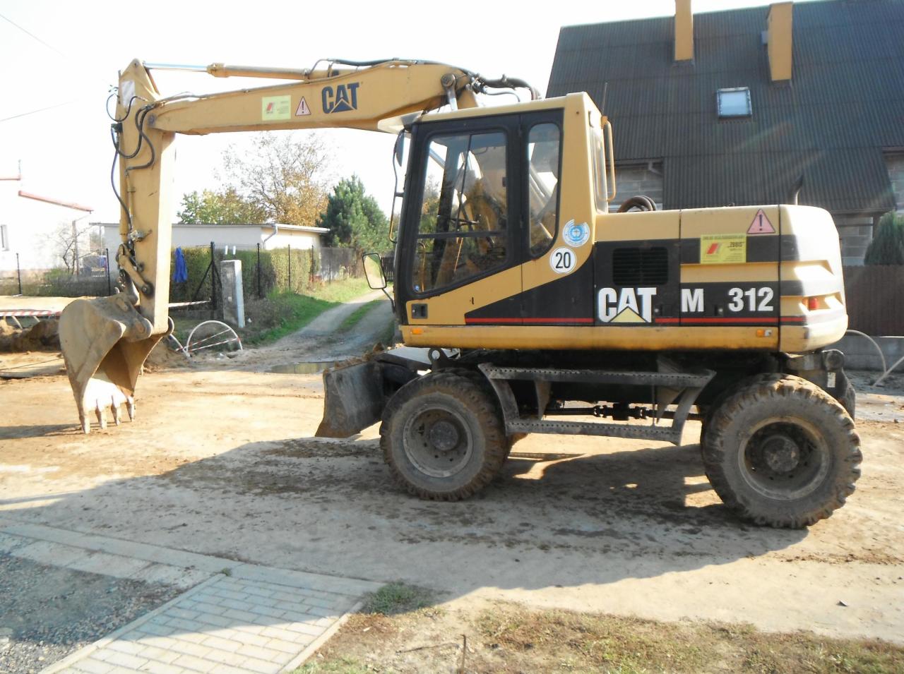Żółta koparka CAT M 312 na placu budowy, z wykopem w tle i widocznymi elementami instalacji.