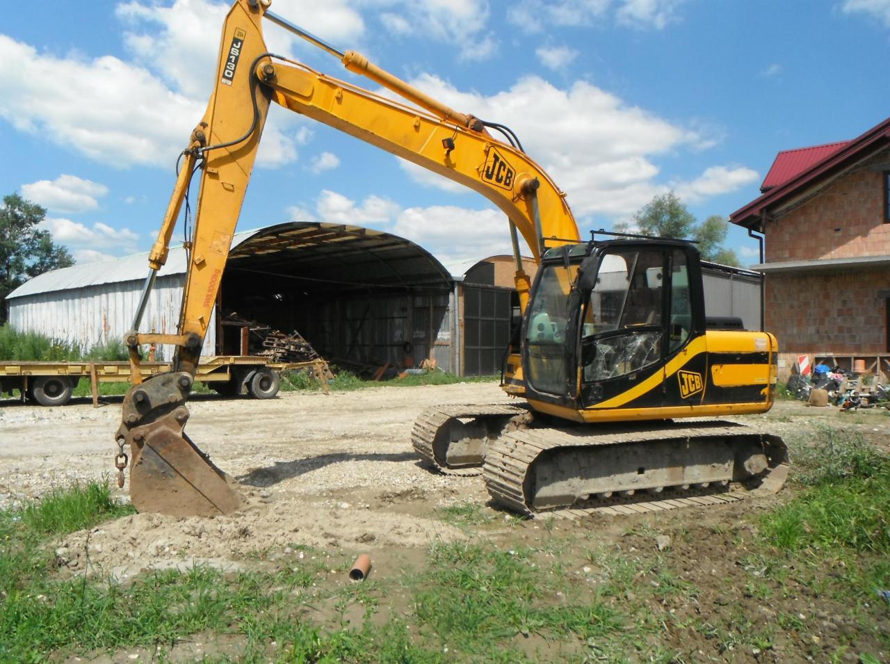 Żółto-czarna koparka gąsienicowa JCB z uszkodzoną szybą kabiny, stojąca na ziemi z widocznym wykopem i rurą PCV, w tle magazyn i budynek w trakcie budowy, błękitne niebo z chmurami.