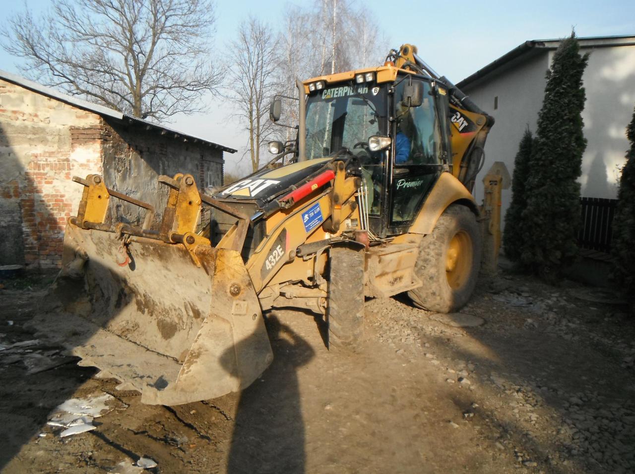Żółta koparko-ładowarka Caterpillar 432E stoi na nierównym terenie z gruzem, obok ceglanego budynku i wysokich tuj, widoczne ślady użytkowania i zabrudzenia na maszynie.