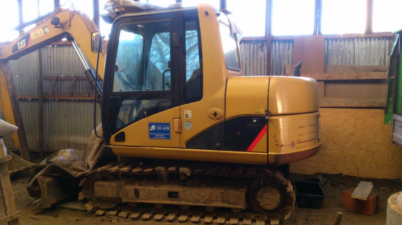 Żółta minikoparka CAT zaparkowana wewnątrz budynku o ścianach z blachy falistej i desek. Na boku koparki widoczna naklejka z danymi firmy.