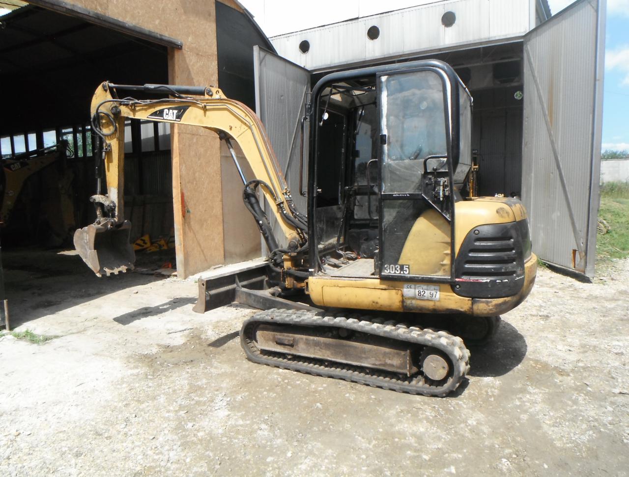 Żółto-czarna mini koparka Caterpillar 303.5 zaparkowana przed otwartym garażem z widoczną inną maszyną w tle, na ziemi drobny żwir.