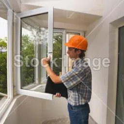 Pracownik w kasku montuje białe okno na balkonie. Widoczne drzewa w tle, częściowo otwarte okno i inne okno w ścianie. Ujęcie z bliska, jasne światło.