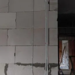 Białe przewody elektryczne prowadzone po ścianie z bloczków betonowych w surowym stanie, widoczne mocowania przewodów, w tle fragment drabiny i otwór drzwiowy.