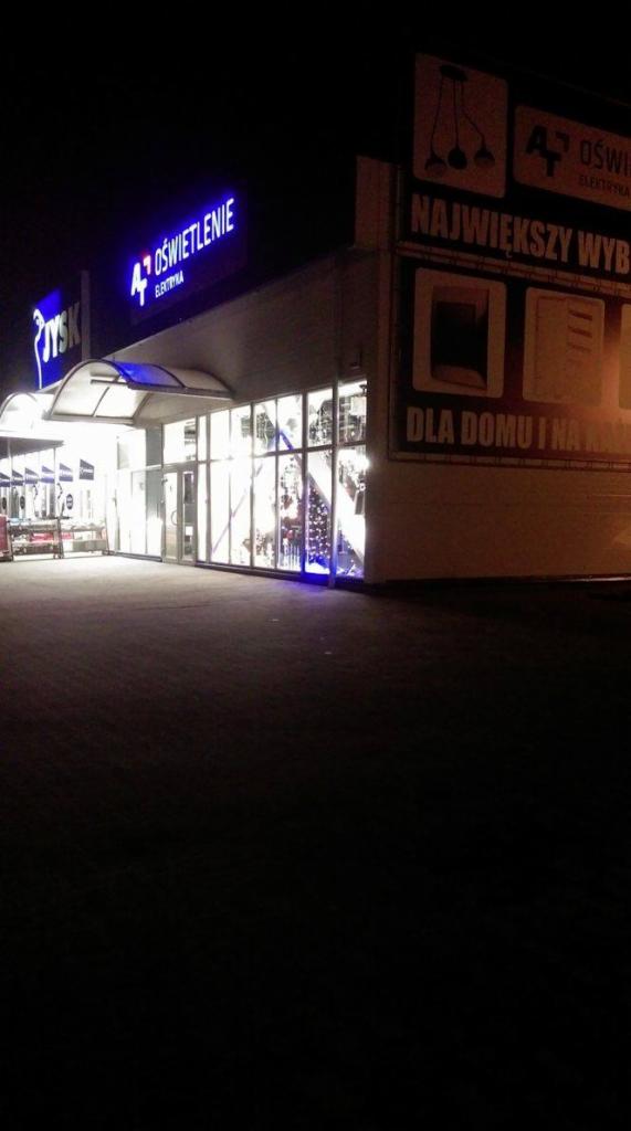 Zewnętrzny widok na sklep z oświetleniem nocą, z widocznymi neonowymi szyldami i wystawą oświetlenia w oknach.