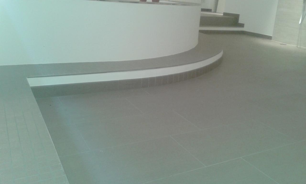 Szare płytki podłogowe ułożone w nowoczesnym, zakrzywionym wnętrzu z białymi ścianami i schodami w tle. Minimalistyczny design z dbałością o detale.