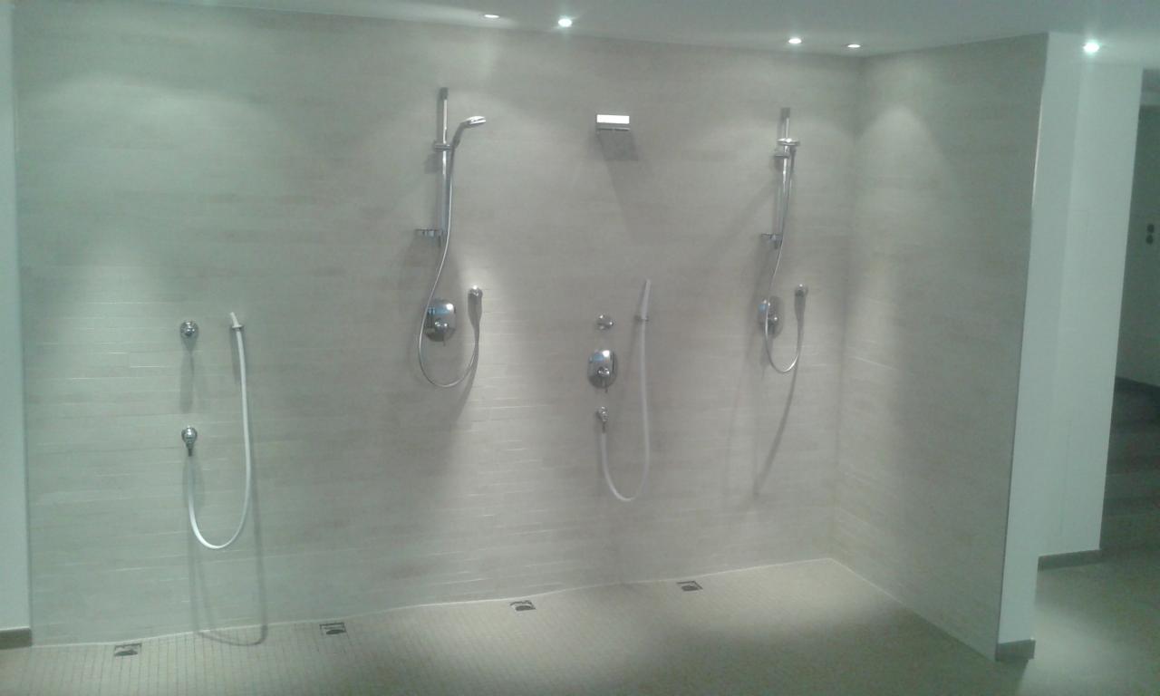 Jasna, wykafelkowana ściana z trzema zestawami prysznicowymi; widoczne baterie, słuchawki i węże. Minimalistyczny design, oświetlenie punktowe.