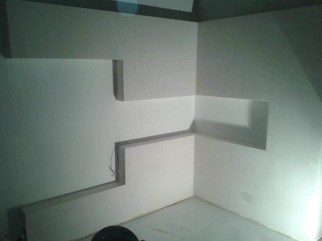 Narożnik pokoju z nietypową, kaskadową zabudową ścian, widoczne przewody. Ściany pomalowane na biało. Częściowo widoczny element wyposażenia na dole.