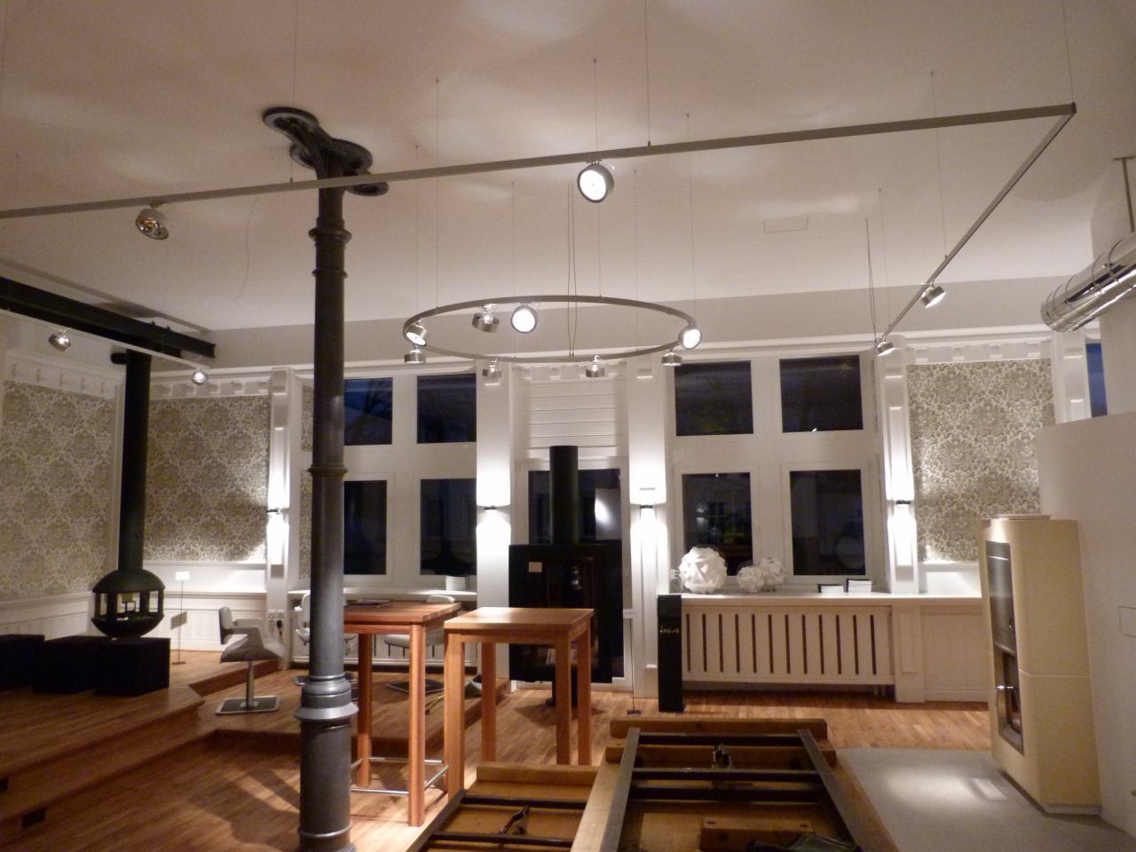 Salon z parkietem, piecem wolnostojącym i oknami w białych ramach. Oświetlenie punktowe i dekoracyjne tapety. Elementy w trakcie montażu na pierwszym planie.