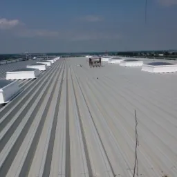 Szary dach z blachy trapezowej z zamontowanymi świetlikami dachowymi, widoczny dźwig w tle.