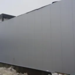 Szara ściana budynku wykonana z płyt warstwowych, widoczne łączenia płyt, w tle budynek w trakcie budowy i śnieg na ziemi.