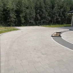 Wyłożony jasnoszarą kostką brukową podjazd do garażu z dekoracyjnymi obrzeżami z ciemniejszej kostki, widok z poziomu gruntu na tle zieleni.