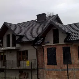 Dom w stanie surowym z nowym, ciemnym pokryciem dachowym, widoczne rynny, komin i drabina oparta o ścianę.